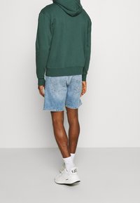 Felpa verde con cappuccio, polsini e orlo a coste, abbinata a pantaloncini di jeans azzurri e sneakers bianche con dettagli neri.