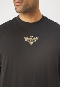 Czarna bawełniana koszulka z długim rękawem z haftowanym złotym logo z skrzydłami na piersi. Okrągły dekolt, gładka tekstura i klasyczny krój.