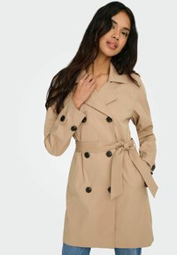 Femme aux longs cheveux bruns portant un trench-coat beige croisé ceinturé à la taille et un jean bleu, debout devant un fond uni.