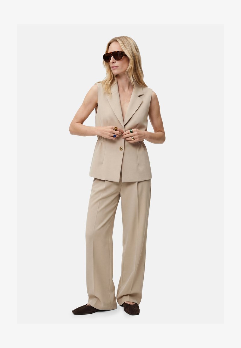Mujer con blazer beige sin mangas y pantalones de pierna ancha, zapatos marrón oscuro, gafas de sol y anillos coloridos, ajustando los botones del blazer.