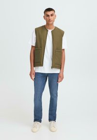 Olijfgroene mouwloze vest met twee voorzakken, gecombineerd met een witte t-shirt en blauwe jeans. Schoeisel is lichtgekleurde sneakers.