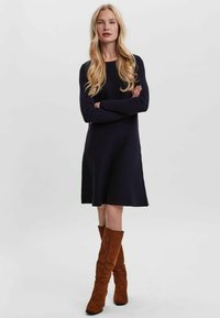 Robe en tricot marine à manches longues, avec un ourlet évasé, portée avec des bottes en daim marron jusqu'aux genoux. Texture lisse, silhouette décontractée.