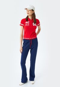 Rode polo-shirt met witte accenten en knopen, gecombineerd met marineblauwe joggers met een trekkoord en zijstrepen, witte pet gedragen.