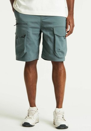 Teal cargo shorts, valmistettu kevyestä kankaasta, joissa on kaksi suurta sivutaskua ja vetoketjudetailit. Yhdistetty vaaleanvärisiin tennareihin.