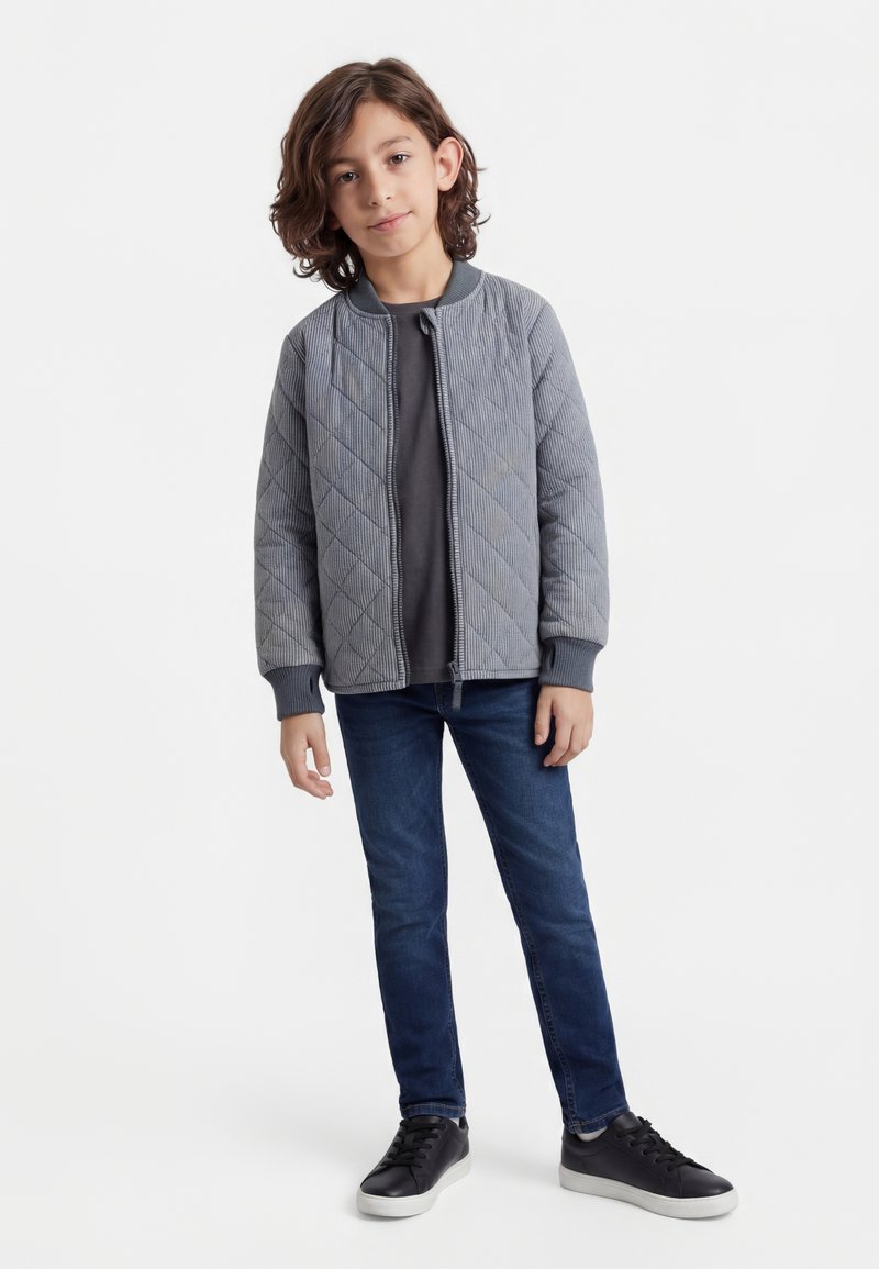 Dreng med skulderlangt brunt hår iført en grå quiltet jakke, mørkegrå skjorte, blå jeans og sorte sneakers, stående mod en hvid baggrund.