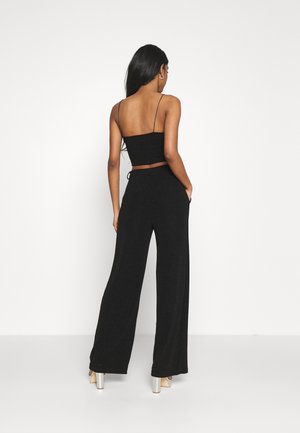 Kvinde med langt sort hår iført sort spaghetti-strop crop top og vide sorte bukser, stående med den ene hånd i lommen.