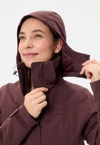 Vrouw die de capuchon van een kastanjebruine Vaude-jack aanpast, met haar gezicht naar links en een lichte glimlach tegen een effen witte achtergrond.