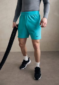 Türkiissinised spordilühikesed püksid tekstureeritud pinnaga, Nike logo, koos mustade tossudega ja valgete pahkluusokkidega. Taust on neutraalne.