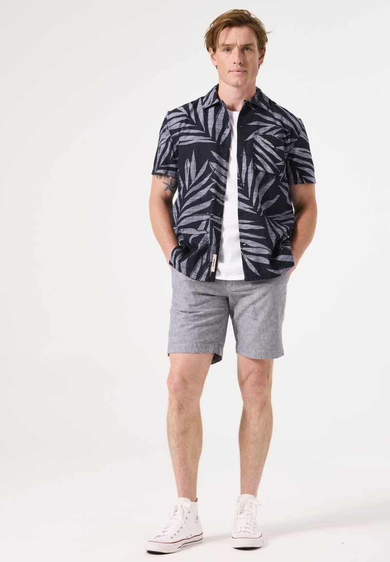 Camisa de manga corta azul marino con patrón de hojas blancas, combinada con pantalones cortos gris claro. Zapatillas deportivas blancas completan el atuendo. Diseño y ajuste casual.