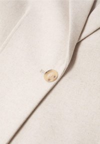 Gros plan sur un tissu beige avec un seul bouton marron clair cousu près d’un bord plié, montrant un détail de manteau ou de veste.