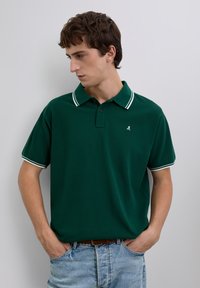 Joven con las manos en los bolsillos, vistiendo una camiseta polo verde oscuro con rayas blancas en el cuello y los bordes de las mangas, mirando hacia su izquierda.