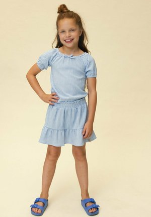 Lachend meisje met lang haar in een blauw kortmouwig topje en rok, met blauwe sandalen, staand met één hand op de heup.