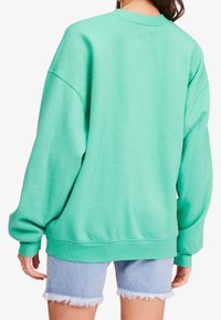 Kvinna i en oversized mintgrön sweatshirt och ljusblå fransiga jeansshorts, sedd bakifrån mot en vit bakgrund.