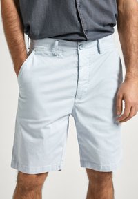 Pantaloni shorts in cotone blu chiaro con fronte piatto, tasche laterali e chiusura a bottone. Taglio dritto con orlo pulito, che mostra una texture minimale.