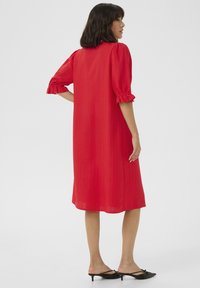 Robe rouge, longueur genou avec des manches bouffantes et des poignets à volants. Texture lisse avec des rayures verticales. Portée avec des mules à talons noirs.