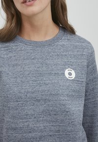 Grå teksturert sweatshirt med en rund hvit knapp med en svart logo på venstre brystområde. Rund halsdesign, middels tungt stoff.