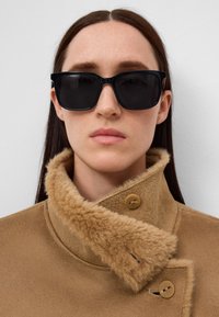Vrouw met steil bruin haar, gekleed in grote zwarte zonnebril en een tan schapenvachtjas die bij de kraag is vastgemaakt tegen een effen achtergrond.