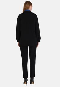 Zwarte oversized jas met lange mouwen en een blauwe kraag, gecombineerd met zwarte maatwerk broek en hoge hakken. Glad textuur, minimalistisch ontwerp.