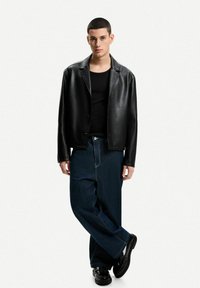 Schwarze Lederjacke mit einfachem Kragen, getragen über einem schwarzen Shirt. Lässige dunkle Jeans und glänzende schwarze Schuhe vervollständigen das Outfit.