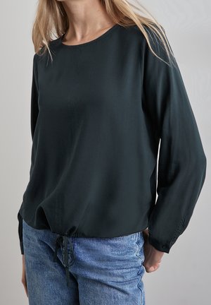 Vrouw draagt een zwarte blouse met lange mouwen, een voornaad en koordje, gecombineerd met een blauwe spijkerbroek, staand tegen een effen achtergrond.