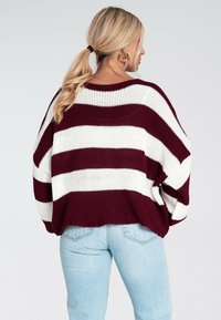 Kobieta z blond włosami związanymi w koński ogon, nosząca złote kolczyki w kształcie koła oraz sweter w biało-bordowe paski, w jasnoniebieskich dżinsach, odwrócona plecami.