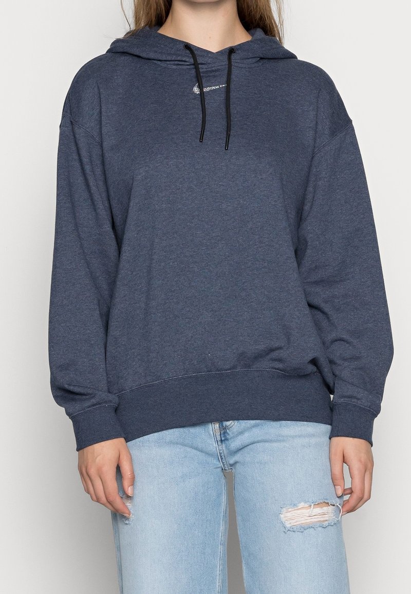 Sudadera azul oscuro con bolsillo tipo canguro frontal, cordón ajustable y puños de canalé. Combinada con jeans claros rotos.