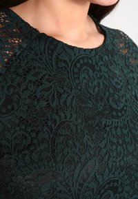 Robe en dentelle vert foncé avec des motifs floraux intricats, un col rond et des détails transparents aux épaules. Tissu texturé avec une doublure lisse.