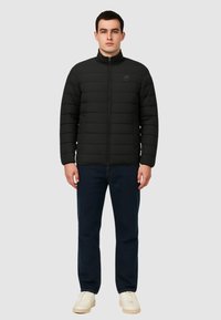 Giacca imbottita nera con colletto alto e zip frontale, caratterizzata da dettagli delle cuciture e un logo sul petto. Abbinata a jeans scuri e scarpe beige.
