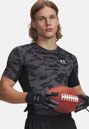 Giovane uomo con maglietta sportiva mimetica nera e grigia che tiene in mano un pallone da football americano con guanti neri Under Armour su sfondo neutro.