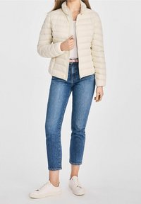 Kobieta ubrana w kremową kurtkę puchową, niebieskie skrócone dżinsy, jasnoróżowy pasek i białe trampki na białym tle.
