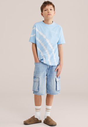 Fant stoji in nosi svetlo modro majico z batik vzorcem, jeans kratke hlače do kolen z žepi, bele nogavice s bež progami in rjave copate brez vezalk.