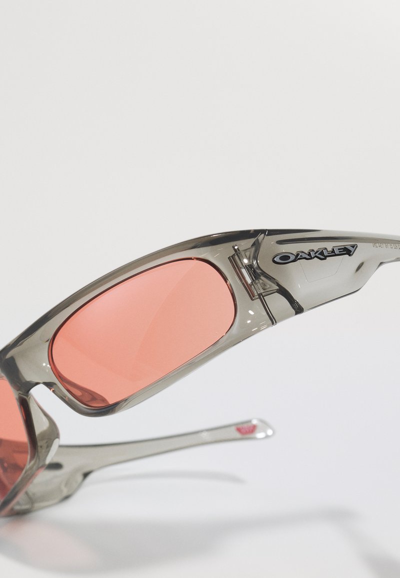 Occhiali da sole Oakley trasparenti grigi con lenti rettangolari rosa su uno sfondo bianco.