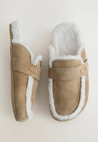 Tan suède slippers met witte nepbont voering, voorzien van een band met een metalen accent gemerkt "SELF." Ronde vorm en gevoerde binnenkant.