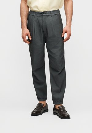 Man draagt donkergrijze taps toelopende broek met elastische boorden, een lichtgele korte mouw shirt en zwarte leren instapschoenen.