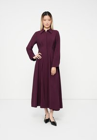 Burgunderfarbenes, knöchellanges Kleid mit durchgehender Knopfleiste, langen Ärmeln und Kragen; hat einen taillierten Oberteil und einen fließenden Rock.