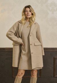 Béžový oversized kabát z texturované látky, s klopovým límcem, dvěma předními kapsami a knoflíkovým zapínáním, nošený přes odpovídající šaty.