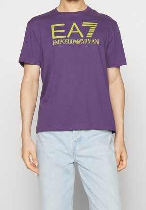 Mężczyzna w fioletowej koszulce z żółtym logo "EA7 Emporio Armani" i jasnoniebieskich spodniach jeansowych, stojący na jednolitym białym tle.