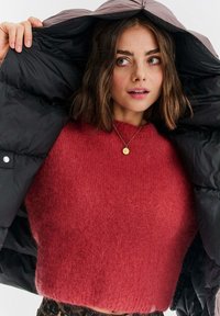 Czerwona, puszysta sweter połączony z czarnym, puchowym płaszczem, z widocznym zamkiem błyskawicznym i detalami w postaci guzików na płaszczu. Złoty naszyjnik z wisiorkiem jako akcent.