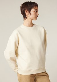 Felpa over-size color crema con colletto a girocollo a costine, spalle scese e texture morbida, abbinata a pantaloni beige.