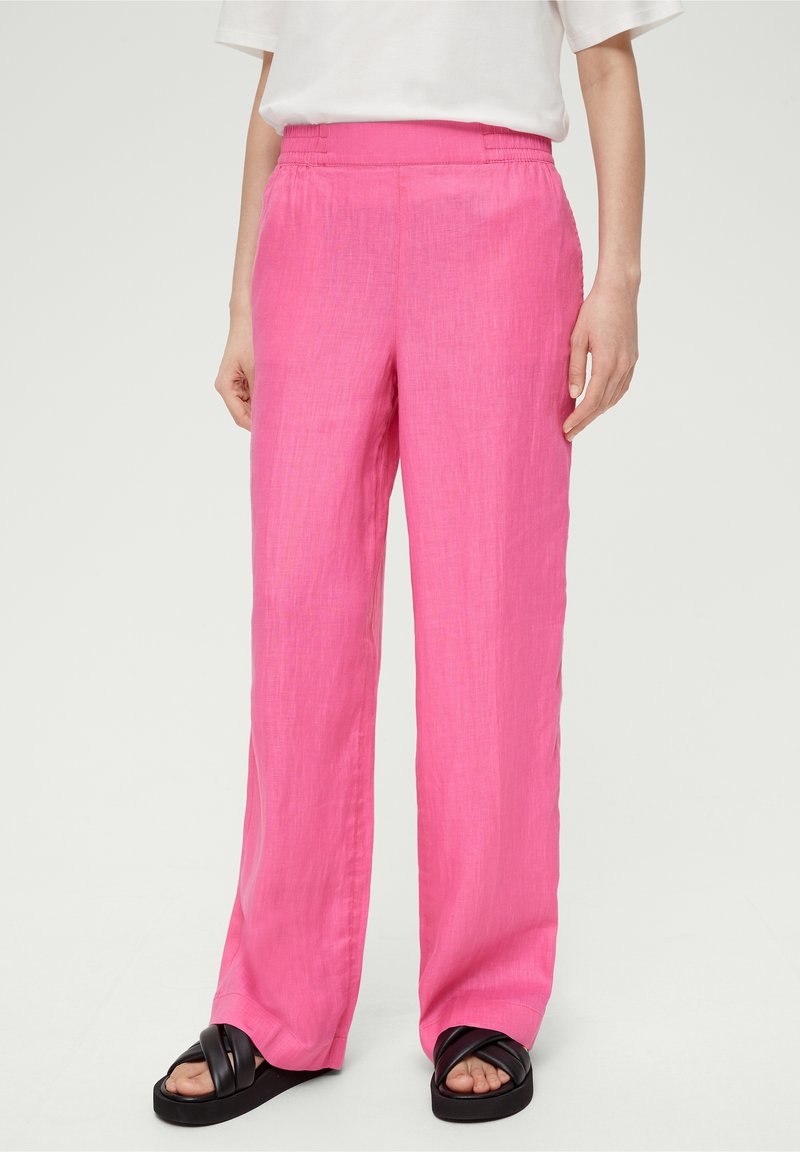 Personne portant un pantalon ample rose vif avec une taille élastique et des sandales noires à bout ouvert sur un fond neutre.