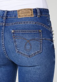 Blue denim jeans with a leather tag reading "KOROSHI." Notable details include a decorative pocket design and pearl accents along the side.