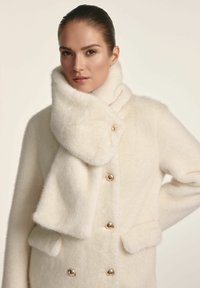 Biały płaszcz z sztucznego futra z wysokim, załamanym kołnierzem i złotymi guzikami. Posiada dwie kieszenie z przodu oraz miękką, puszystą fakturę.