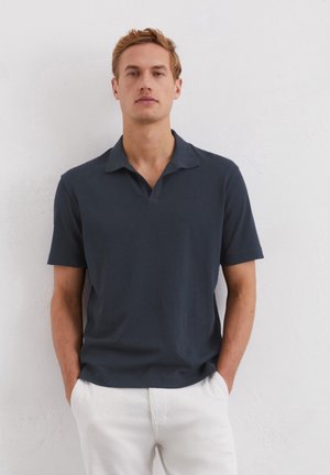 Navyblauwe poloshirt met korte mouwen en een kraag, gemaakt van een gladde stof. Het model staat met de handen in de zakken en draagt lichtgekleurde broek.