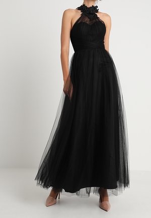 Kvinde iført en ærmeløs sort tyl aftenkjole med blomsterdekoration ved halsudskæringen og nude høje hæle.