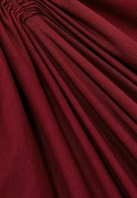 Tessuto rosso scuro con superficie testurizzata, caratterizzato da pieghe verticali che creano un aspetto stratificato. Finitura liscia e leggermente lucida.
