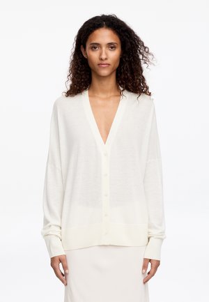 Žena s kučeravými vlasmi má na sebe voľný biely zapínaný sveter a zodpovedajúcu sukňu, stojí na jednoduchom bielom pozadí.