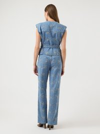 Γυναίκα που στέκεται πλάτη, φορώντας μπλε κεντημένο τζιν γιλέκο και ταιριαστό καμπάνα παντελόνι με ψηλά τακούνια, μπροστά σε απλό φόντο.