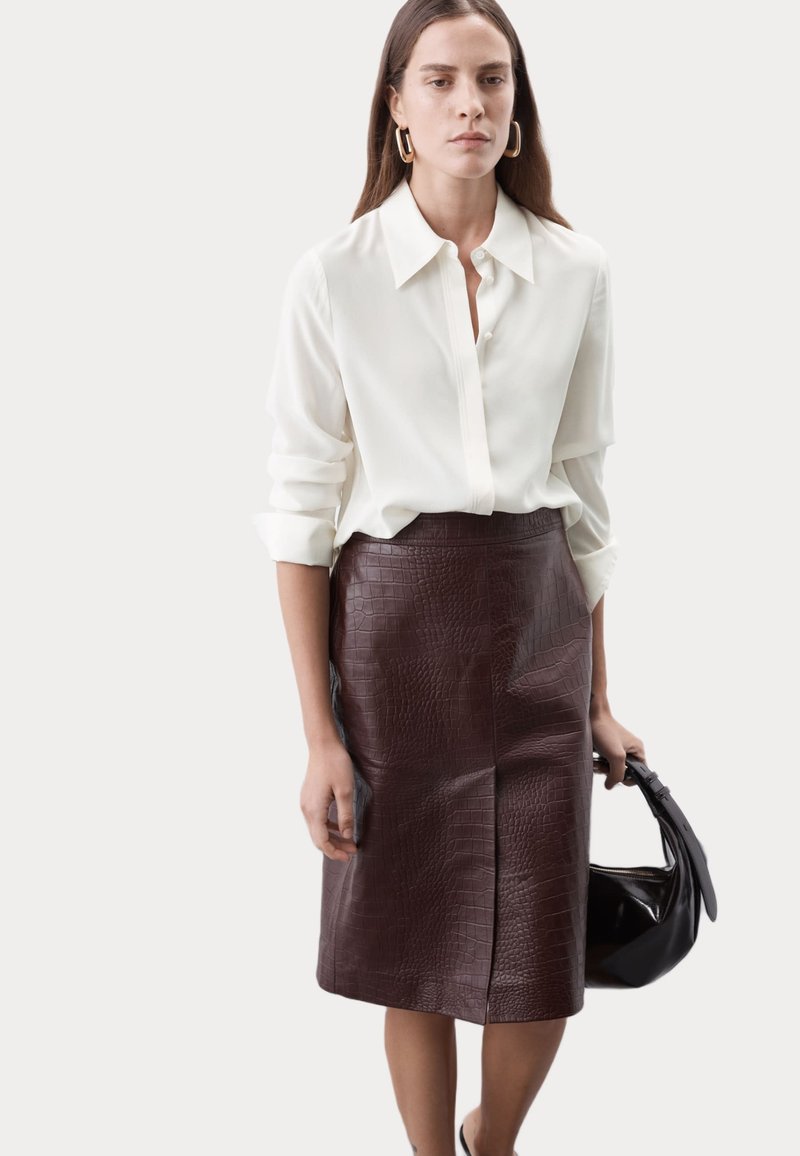 Camisa blanca de manga larga con cuello abotonado combinada con una falda de cuero burdeos de largo hasta la rodilla con relieve de cocodrilo. La modelo sostiene un bolso negro.