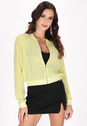 Yellow textured bomber jacket with a zip front, ribbed cuffs, and hem, worn over a black top. Model poses neutrally against a plain background.
