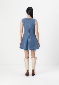Denim mouwloze jurk met een uitlopende rok, voorzien van verticale stiksel en een ritssluiting aan de achterkant. Gecombineerd met knie-hoge crème en bruine laarzen.